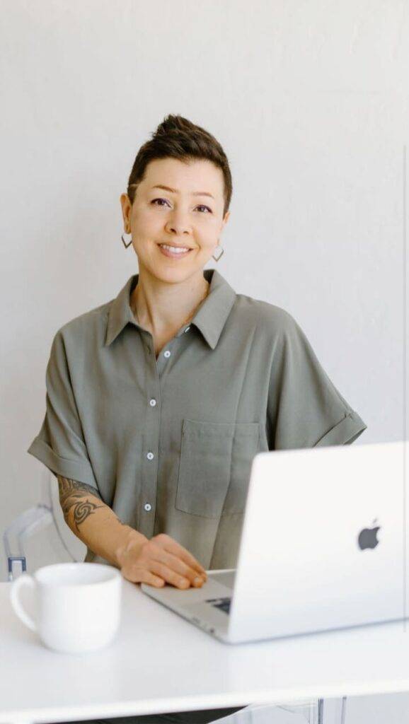 Woman with short hair and a tattooed arm smiling at a desk with a laptop and a coffee mug